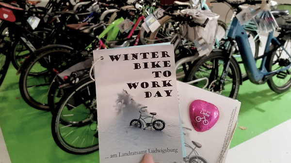Un groupe de vélos alignés dans un espace intérieur. Au premier plan, une affiche sur le "Winter Bike to Work Day" et un autocollant en forme de cœur avec une image de vélo. L'événement est organisé à Ludwigburg.