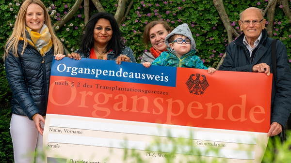 A group of five people stands together outdoors, holding a large, oversized organ donation card that reads "Organspendeausweis." The card includes sections for personal details. They appear to be promoting organ donation awareness. The background features greenery.