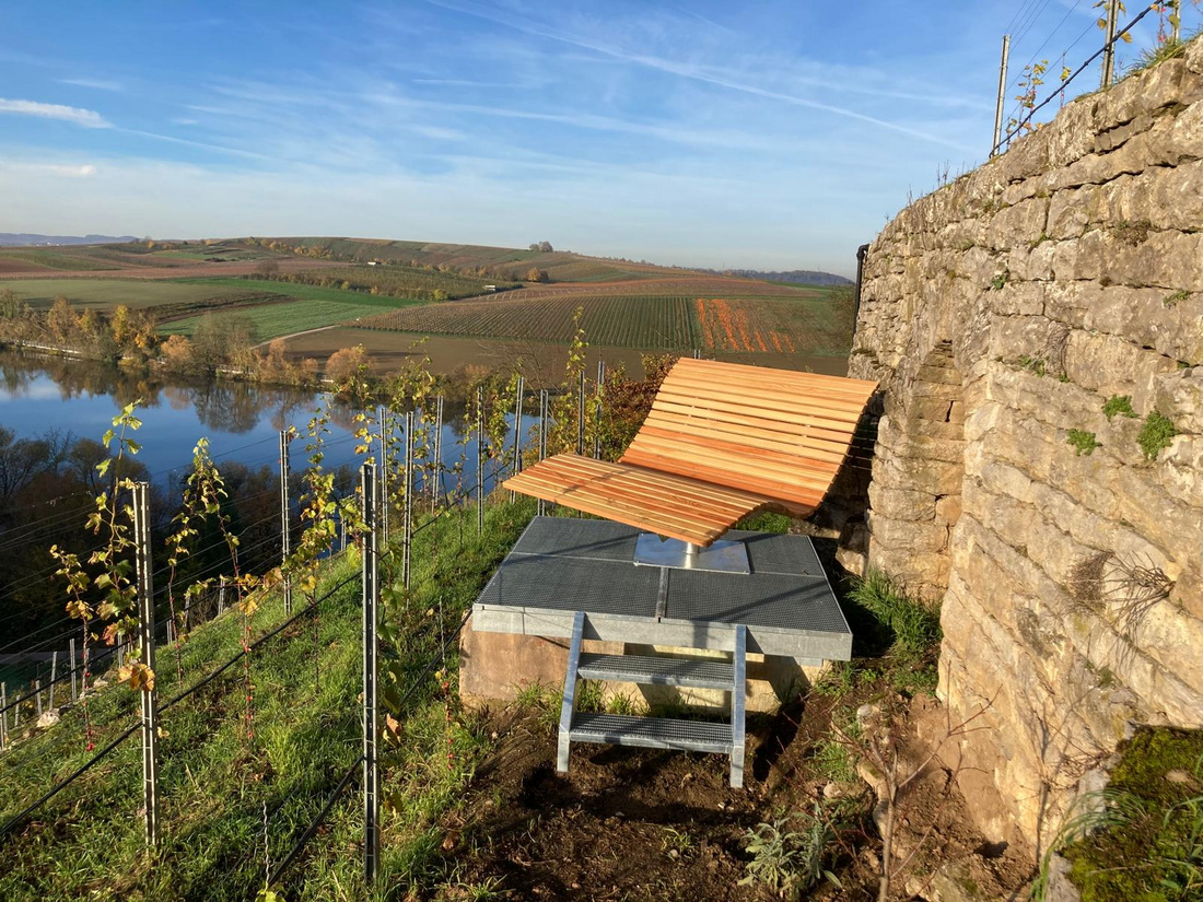 Eine Holzliege steht inmitten von Weinbergen mit Blick auf einen ruhigen Fluss und sanfte Hügel im Hintergrund. Die Umgebung zeigt lebendige grüne Pflanzen und einen klaren blauen Himmel. Die Szene vermittelt ein Gefühl von Ruhe und Naturverbundenheit.