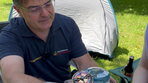 Ein Mann sitzt an einem Tisch im Freien und hält einen Campingkocher in der Hand, während er ihn betrachtet. Neben ihm sind Utensilien und ein Kochtopf zu sehen. Im Hintergrund ist ein Zelt und Bäume zu erkennen.