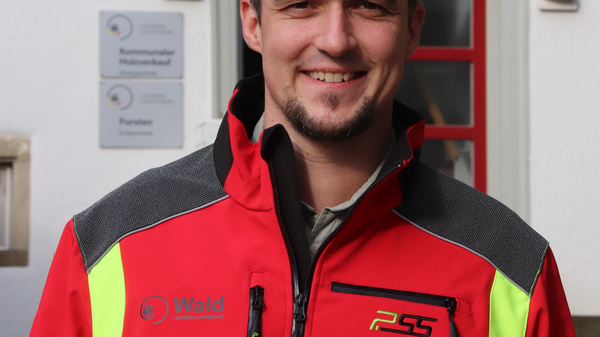 A man with short grey hair and a beard smiles while wearing a bright red jacket with reflective yellow details. He is standing outdoors, in front of a building with a red door and windows.