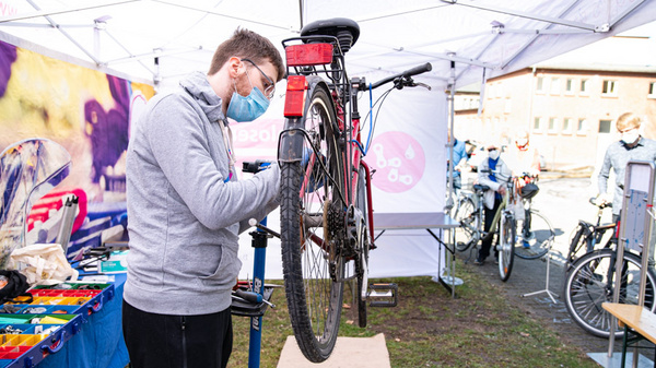 Un homme portant un masque répare un vélo dans un espace extérieur sous une tente. Des outils sont visibles à proximité, et d'autres vélos sont stationnés derrière lui. L'ambiance est celle d'un événement de maintenance de vélos, avec des acteurs et des participants autour.