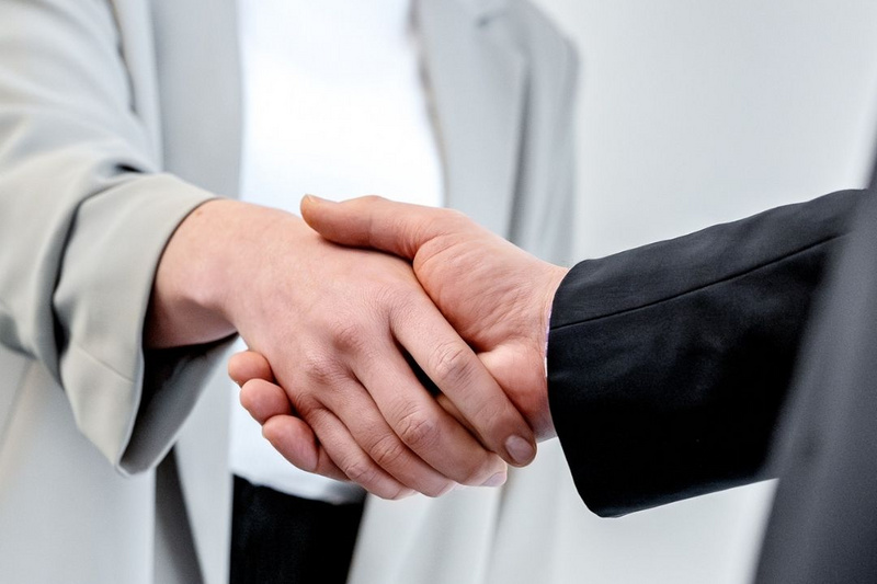 Eine Frau in einem grauen Blazer und ein Mann mit dunklem Anzug schütteln sich die Hände. Die Geste symbolisiert einen Geschäftsabschluss oder eine freundliche Begrüßung.