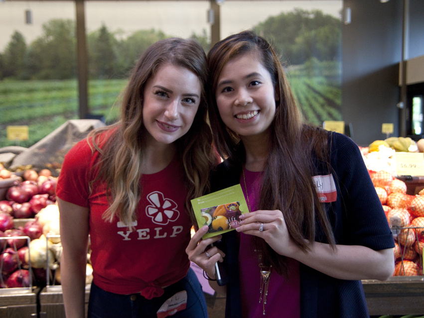 Bild: Elite Sneak Peek of PCC Bothell, Lizenz: CC BY-NC-ND 2.0 Zwei Frauen stehen in einem Lebensmittelgeschäft vor einem Ständer mit Äpfeln. Die Frau links trägt ein rotes T-Shirt mit dem Yelp-Logo. Beide lächeln und halten eine Karte in der Hand. Im Hintergrund sind grüne Felder sichtbar.