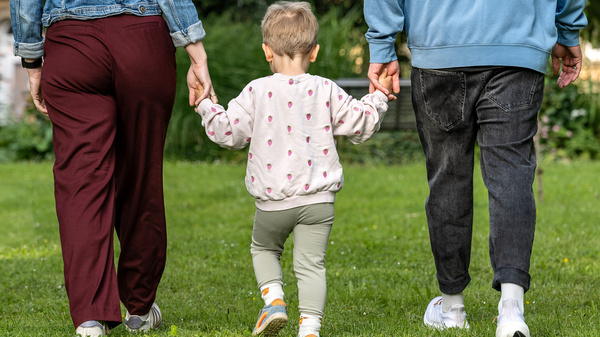 Ein Kind hält die Hände von zwei Erwachsenen und geht auf einer grünen Wiese. Die Person auf der linken Seite trägt eine Jeansjacke und rote Hose, die Person auf der rechten Seite trägt einen blauen Pullover und schwarze Jeans. Das Kind trägt ein gepunktetes Sweatshirt und grüne Hosen.