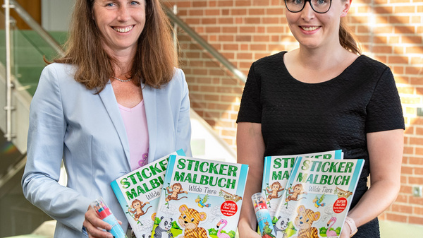 Zwei Frauen stehen im Innenraum, lächelnd und halten jeweils ein Sticker-Malbuch in den Händen. Die Farbgestaltung der Bücher zeigt Tiere, die für Kinder ansprechend sind. Im Hintergrund sind helle Wände und Fenster zu sehen, die eine freundliche Atmosphäre schaffen.