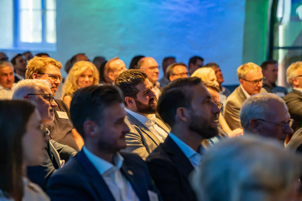 Eine Gruppe von Personen sitzt in einem großen Raum und hört einer Präsentation zu. Die Zuschauer zeigen Interesse und sind in formeller Kleidung. Das Licht im Raum ist sanft und sorgt für eine angenehme Atmosphäre.
