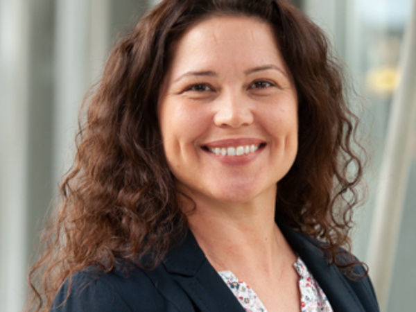 A woman with long, curly dark hair smiles warmly at the camera, wearing a black blazer over a floral blouse. The background is blurred, emphasizing her friendly expression and professional appearance.