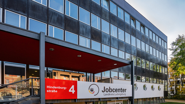 A modern building featuring large windows and a red entryway. A sign prominently displays "Jobcenter" along with the address "Hindenburgstraße 4." The surrounding area includes green landscaping.