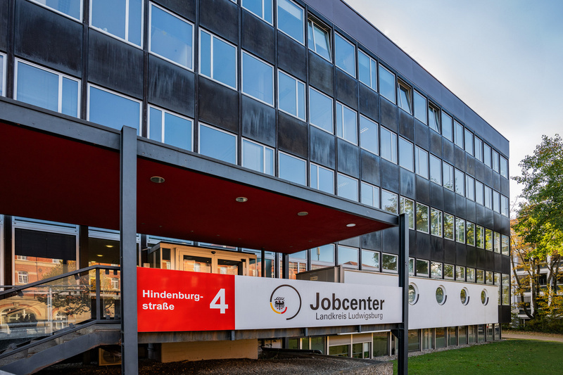 Das Bild zeigt das Gebäude des Jobcenters in der Hindenburgstraße 4. Der Eingang ist mit einer roten Überdachung versehen, und ein Schild mit der Aufschrift „Jobcenter Landkreis Ludwigslust-Parchim“ ist deutlich sichtbar. Das Gebäude hat moderne Fenster und eine gepflegte Umgebung.