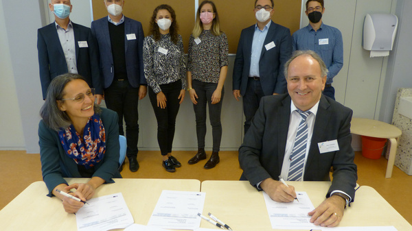 Deux personnes assises à une table signent des documents. En arrière-plan, un groupe de sept personnes observe, portant des masques faciaux. L'ambiance semble professionnelle et formelle, suggérant un événement ou un accord important.