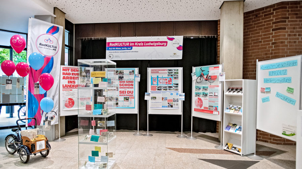 Un stand d'exposition présentant des informations sur le projet "Radkultur" dans le district de Ludwigsbourg. Des panneaux explicatifs, des brochures et un vélo sont visibles. Des ballons colorés ajoutent une touche festive à l'événement.
