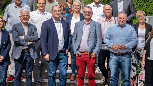 Eine Gruppe von 15 Personen steht im Freien vor einer grünen Kulisse. Die Teilnehmer sind unterschiedlich gekleidet und lächeln. Sie repräsentieren eine vielfältige Gemeinschaft, die zusammengekommen ist, wahrscheinlich für ein Treffen oder ein Event.
