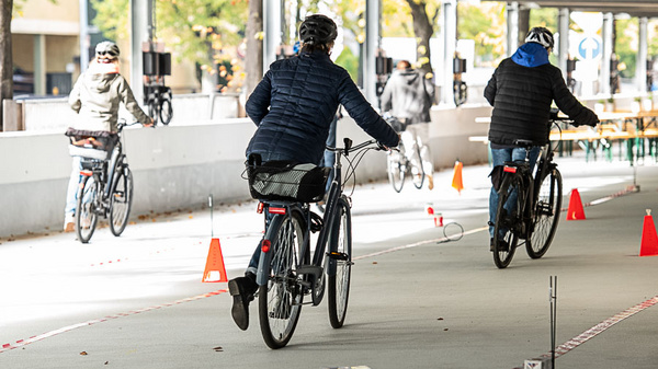Drei Fahrradfahrer in leichter sportlicher Kleidung fahren auf einem befestigten Weg. Im Hintergrund sind Verkehrszeichen und Hindernisse in Form von roten Pylonen zu sehen, die auf eine Fahrradübung oder -schulung hinweisen. Die Umgebung zeigt eine begrünte Fläche.