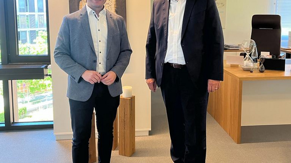 Zwei Männer stehen in einem modernen Büro. Der linke Mann trägt eine graue Jacke und Sneakers, der rechte Mann einen dunklen Anzug und eleganten Schuhen. Beide lächeln und stehen neben einem Tisch mit Büromaterial. Im Hintergrund ist ein Fenster mit natürlichem Licht sichtbar.