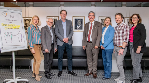 Eine Gruppe von sieben Personen steht in einem Bürogebäude. Sie lächeln und posieren für ein Foto. Im Hintergrund sind Rahmen mit Porträts an der Wand zu sehen. Auf einer Flipchart steht eine Grafik mit Strichmännchen. Die Szene vermittelt eine freundliche, professionelle Atmosphäre.