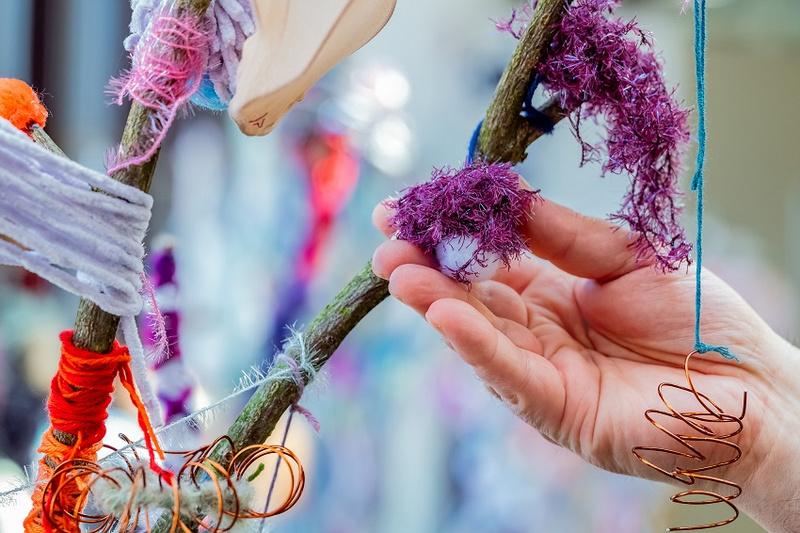 Eine Hand berührt einen verzweigten Ast, der mit bunten Garntüchern und Drahtspiralen dekoriert ist. Die Texturen wirken weich und verschlungen, während die Farben lebhaft und ansprechend sind. Der Fokus liegt auf der Interaktion zwischen der Hand und dem kunstvoll gestalteten Ast.