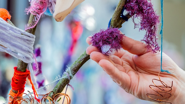 Eine Hand berührt einen verzweigten Ast, der mit bunten Garntüchern und Drahtspiralen dekoriert ist. Die Texturen wirken weich und verschlungen, während die Farben lebhaft und ansprechend sind. Der Fokus liegt auf der Interaktion zwischen der Hand und dem kunstvoll gestalteten Ast.