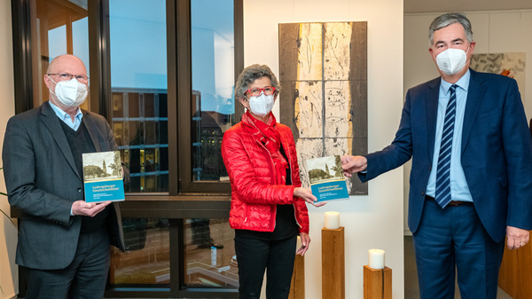 Drei Personen stehen in einem Raum und halten Bücher in den Händen. Die Frau in der Mitte trägt eine rote Jacke und eine Brille. Alle drei Personen tragen Gesichtsmasken. Im Hintergrund sind Fenster und dekorative Elemente zu sehen. (Text KI generiert)