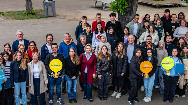 Eine große Gruppe von Menschen steht auf einem Platz und lächelt in die Kamera. Viele halten bunte Schilder in verschiedenen Farben, die möglicherweise Botschaften oder Themen darstellen. Im Hintergrund sind Bäume und Gebäudefassade sichtbar.