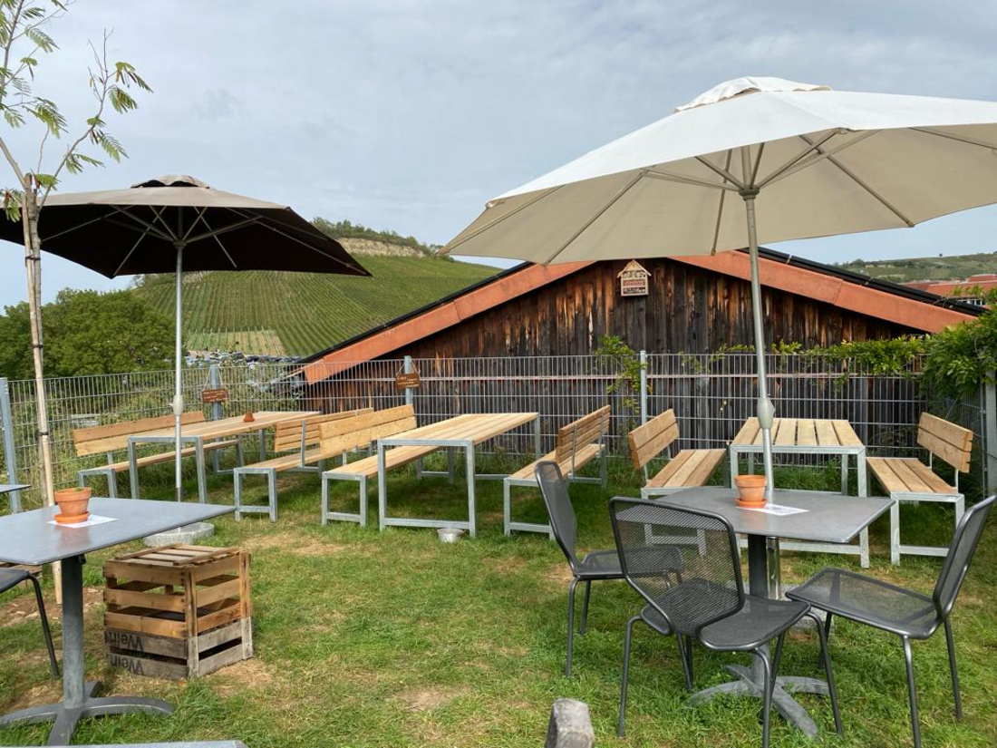 Ein Außenbereich eines Lokals mit Möbeln aus Metall und Holz. Es sind mehrere Tisch- und Bankkombinationen unter sonnenschützenden Schirmen angeordnet. Im Hintergrund ist ein Weinberg und ein rustikales Holzgebäude zu sehen, das die ländliche Atmosphäre unterstützt.