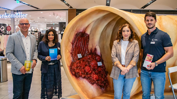 Four individuals stand in a shopping center near a large, artistic representation of an anatomical structure, possibly a gastrointestinal model. They are smiling and holding brochures, promoting health or educational information related to the installation. The environment appears modern and well-lit.