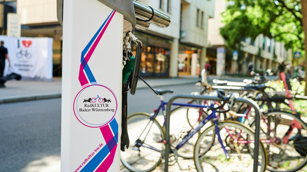 A bike-sharing station is featured prominently, displaying a colorful logo for "RadKULTUR Baden-Württemberg." In the background, several bicycles are parked, and a tree-lined street can be seen, indicating a vibrant urban environment focused on cycling.