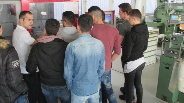 A diverse group of individuals attentively listens to an instructor in a workshop setting. The instructor stands in front of a machine, explaining its operation, while students are gathered closely around, engaged in the discussion. Industrial equipment is visible in the background.
