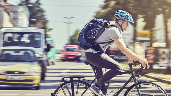 A cyclist wearing a blue helmet and a backpack rides on a street. The background features blurred vehicles and trees, indicating a busy urban setting. The cyclist is focused on the road ahead, contributing to the dynamic atmosphere of city life.