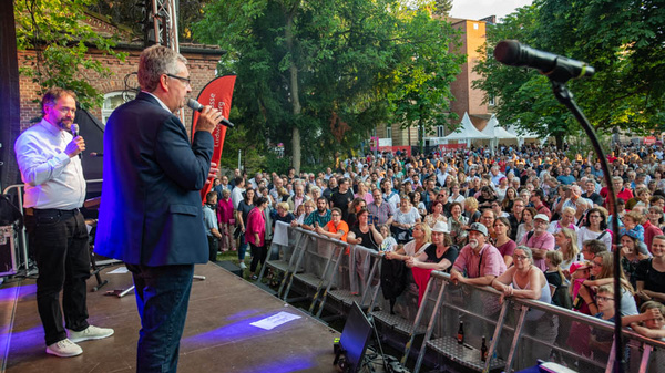 Due relatori si esibiscono su un palco di fronte a una grande folla durante un evento all'aperto. Gli spettatori, visibilmente coinvolti, ascoltano attentamente mentre la luce solare filtra attraverso gli alberi circostanti. Microfoni e attrezzature sono visibili, contribuendo all'atmosfera di festa.