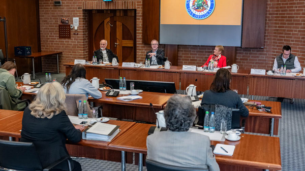 Das Bild zeigt eine Sitzung im Landratsamt Ludwigsburg. An einem großen Tisch sitzen mehrere Personen, während im Hintergrund das Wappen von Ludwigsburg auf einer Leinwand zu sehen ist. Die Anwesenden diskutieren ernsthaft, einige machen sich Notizen.