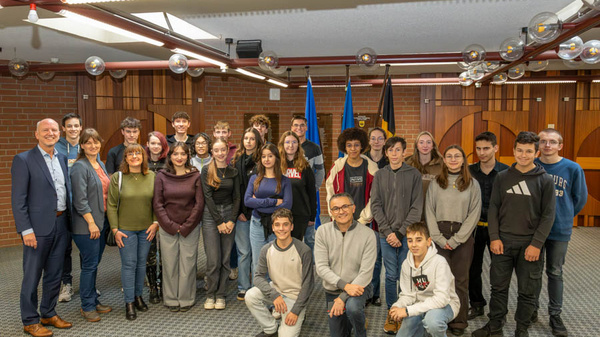 Eine Gruppe von 23 Personen steht in einem Raum, darunter Jugendliche und Erwachsene. Im Hintergrund sind mehrere Flaggen sichtbar. Die Anwesenden lächeln und posieren gemeinsam für das Foto. Der Raum hat eine moderne Gestaltung mit einer modernen Decke und heller Beleuchtung.