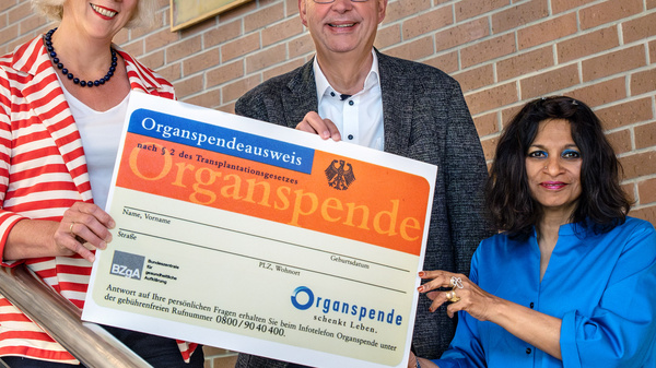 Drei Personen stehen an einer Treppe und halten gemeinsam einen Organspendeausweis hoch. Die Person links trägt eine gestreifte Bluse, die in der Mitte eine Brille und einen Anzug, und die Person rechts ein blaues Kleid. Sie zeigen Engagement für die Organspende.
