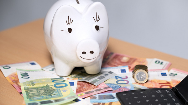 Ein Sparschwein aus Keramik steht auf einem Tisch, umgeben von verschiedenen Geldscheinen in Euro, darunter 20- und 100-Euro-Scheine. Neben dem Sparschwein liegt eine Münze und eine schwarze Geldbörse. Die Szene vermittelt das Thema Sparen und Geldverwaltung.
