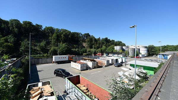 Ein großer Parkplatz mit mehreren Containern für Abfälle, umgeben von Bäumen. Im Hintergrund sind einige Lagerbehälter sichtbar. Der Himmel ist klar und blau, es ist Tageslicht. Ein Auto steht auf dem Platz, während die Umgebung ruhig wirkt.