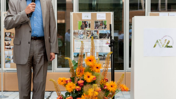 A man in a gray suit stands on a stage, holding a microphone and speaking to an audience. In the background, there are photo displays, and in front of him, a floral arrangement with orange flowers is visible. A podium with a logo is positioned beside him.