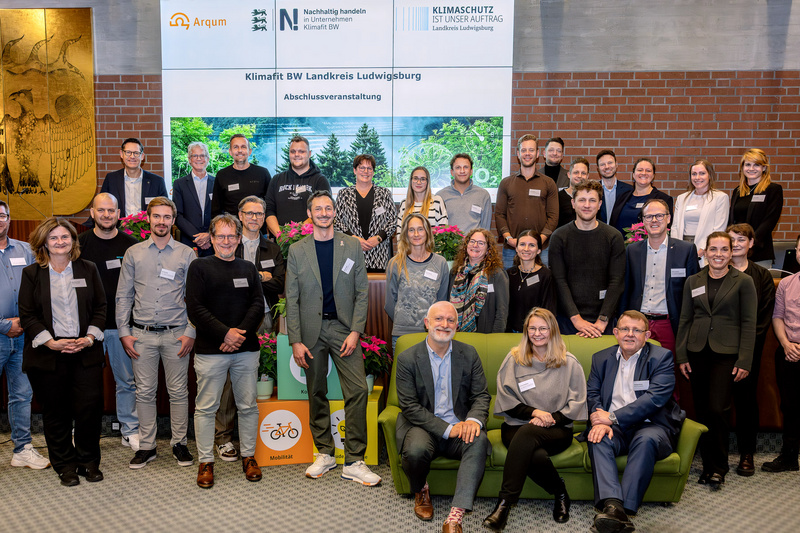 Eine Gruppe von etwa 30 Menschen steht und sitzt in einem Raum, um ein Ereignis zu feiern. Sie tragen Namensschilder und lächeln in die Kamera. Im Hintergrund befindet sich ein Banner mit Informationen zu Klimaschutz und einem Logo.