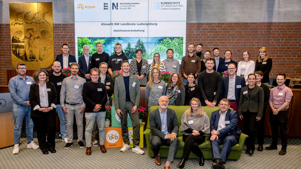 Eine Gruppe von etwa 30 Menschen steht und sitzt in einem Raum, um ein Ereignis zu feiern. Sie tragen Namensschilder und lächeln in die Kamera. Im Hintergrund befindet sich ein Banner mit Informationen zu Klimaschutz und einem Logo.