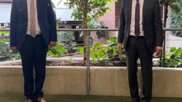 Deux hommes en costume se tiennent debout dans un espace intérieur avec des plantes vertes en arrière-plan. L'un porte un costume bleu et cravate, l'autre un costume noir avec une chemise blanche. Ils sourient et semblent être engagés dans une conversation amicale.