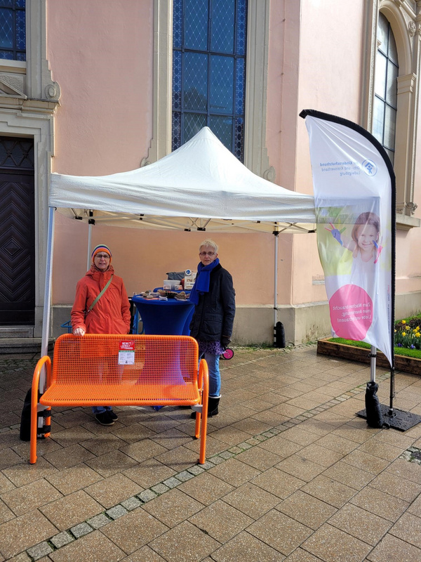 Due donne sorridenti si trovano davanti a un gazebo bianco, una indossa un cappotto arancione e l'altra un cappotto nero. Accanto a loro c'è una panchina arancione e un tavolo con materiale informativo. Sullo sfondo si intravede una parete rosa.