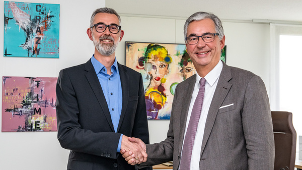 Zwei Männer in Anzügen schütteln sich die Hände in einem Büro. Im Hintergrund sind bunte Kunstwerke an der Wand zu sehen. Die Szene vermittelt einen formellen, geschäftlichen Austausch oder eine Vereinbarung.