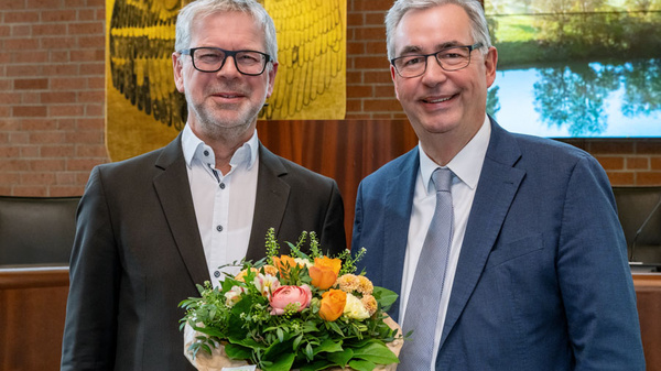 Zwei Männer stehen lächelnd in einem offiziellen Raum. Der Mann links trägt eine Brille und hält einen bunten Blumenstrauß. Der Mann rechts trägt einen blauen Anzug. Im Hintergrund ist eine Wand mit einem Wappen und einem Bild von Landschaften sichtbar.