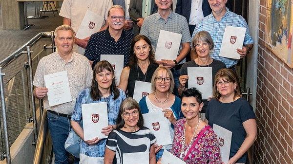 Eine Gruppe von 15 Personen steht in einem Foyer und hält Urkunden in den Händen. Die Mehrheit ist lächelnd und trägt unterschiedliche Kleidung. Im Hintergrund sind Wände und verschiedene Beleuchtungselemente zu sehen. Die Stimmung wirkt feierlich, wahrscheinlich in einem festlichen Kontext.