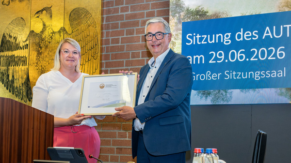 Die Abbildung zeigt eine Frau und einen Mann, die sich in einem Sitzungssaal gegenüberstehen. Der Mann überreicht der Frau ein Zertifikat oder eine Auszeichnung. Im Hintergrund ist ein Banner mit dem Datum und dem Titel der Sitzung zu sehen.