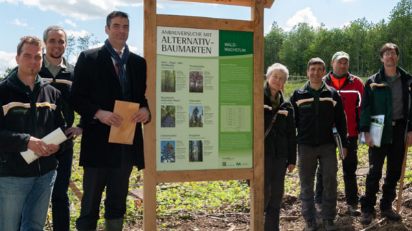 A group of eight people stands together outdoors near a large informational sign about an alternative forestry initiative. The setting appears to be a cleared forest area, with trees in the background and a partly cloudy sky above.
