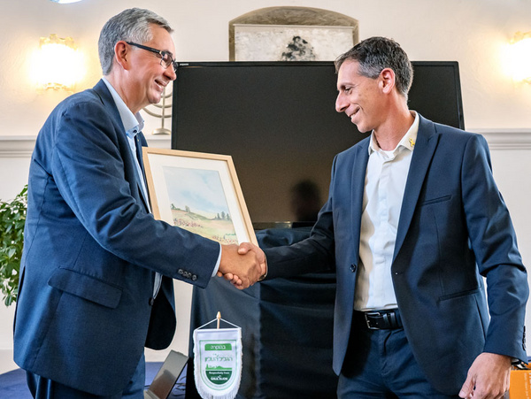 29.05.2025 - Begrüßung des neuen Chairman Asaf Langleben - Oberes Galiläa im Pädagogisch Kulturellen Centrum Zwei Männer schütteln sich die Hände, während einer von ihnen ein gerahmtes Bild überreicht. Im Hintergrund ist ein Bildschirm und eine Pflanze sichtbar. Die Szene deutet auf eine formelle Anerkennung oder Auszeichnung hin.