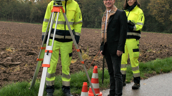 Drei Personen stehen auf einem Feldweg. Zwei in auffälligen, gelben Arbeitsanzügen halten ein Vermessungsgerät, während der dritte Mann in einem dunklen Anzug daneben steht. Orangefarbene Verkehrshütchen sind im Vordergrund sichtbar. Bäume im Hintergrund zeigen die Herbstsaison.