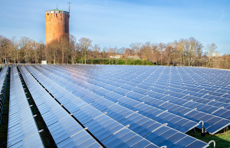 Eine große Photovoltaikanlage mit vielen Solarzellen erstreckt sich über das Bild. Im Hintergrund steht ein runder, roter Wasserturm zwischen Bäumen, unter einem klaren blauen Himmel. Die Szene verdeutlicht den Einsatz erneuerbarer Energien in einer urbanen Umgebung.