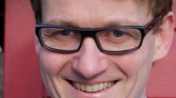 Ein Mann mit kurzem, hellbraunem Haar und Brille lächelt freundlich in die Kamera. Er trägt ein hellblaues Hemd und steht vor einer roten Wand.