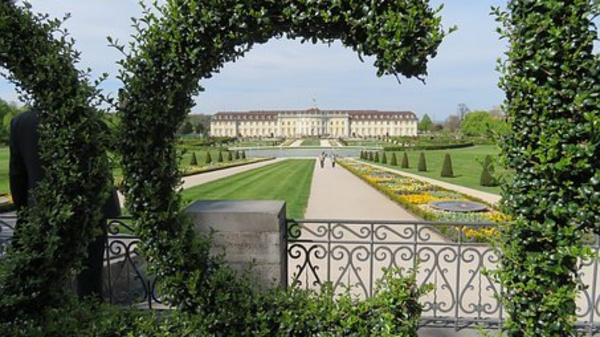 A landscaped garden features neatly trimmed hedges and flowerbeds, leading to a grand building in the background. The path is flanked by geometric shaped trees, creating a symmetrical, tranquil atmosphere.